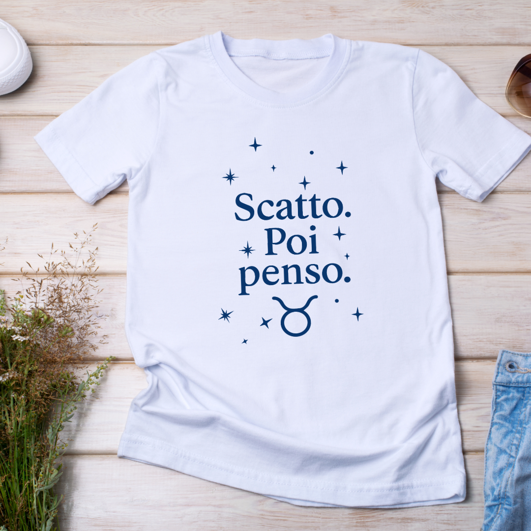 flat lay su legno chiaro):
Maglietta bianca piegata con scritta "Scatto. Poi penso." e simbolo del Toro, decorata con stelline, fotografata dall’alto su fondo in legno chiaro.