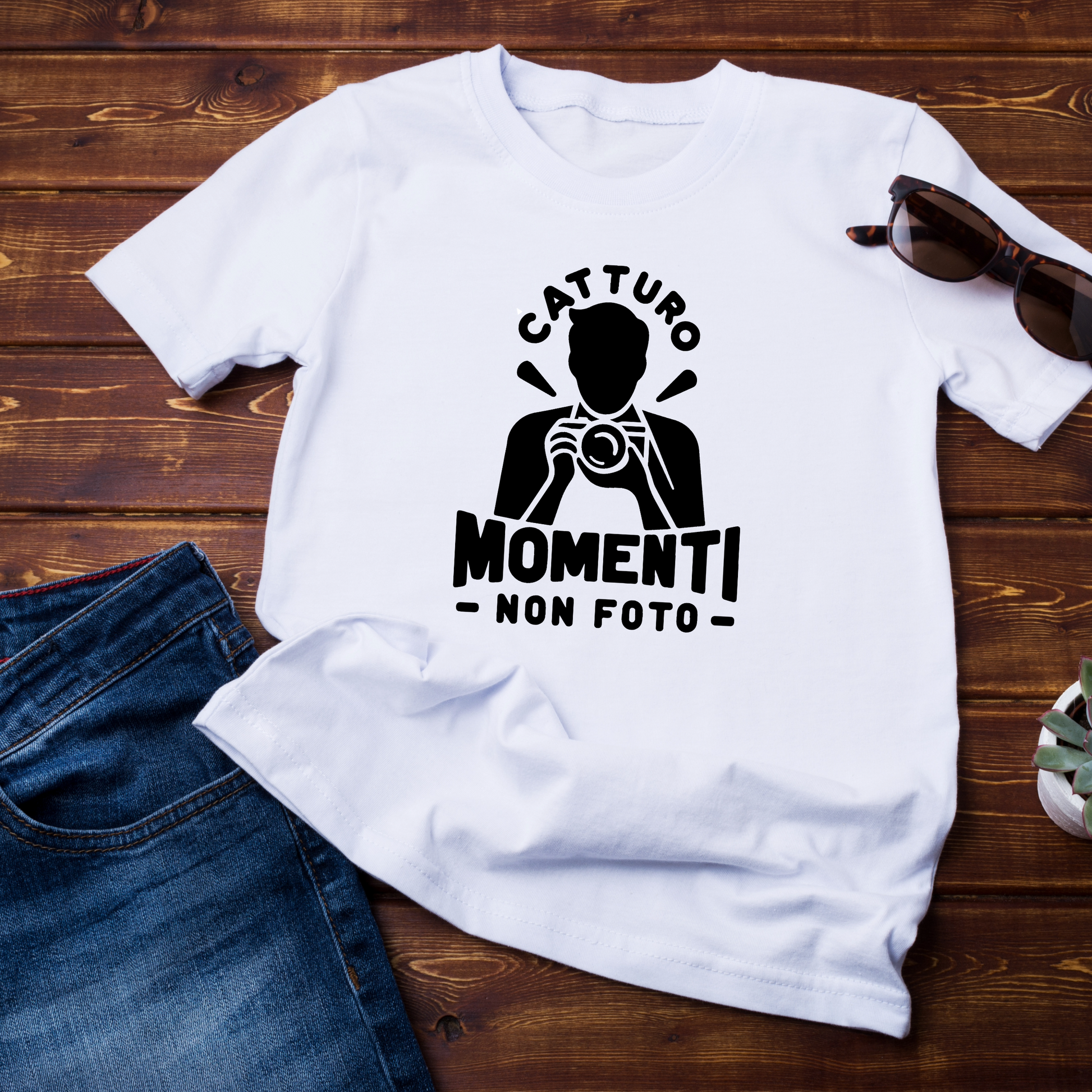 "T-shirt bianca con stampa minimalista in nero che raffigura una silhouette di fotografo con la scritta 'Catturo momenti, non foto'. Disposta su fondo in legno con jeans e occhiali da sole come accessori."