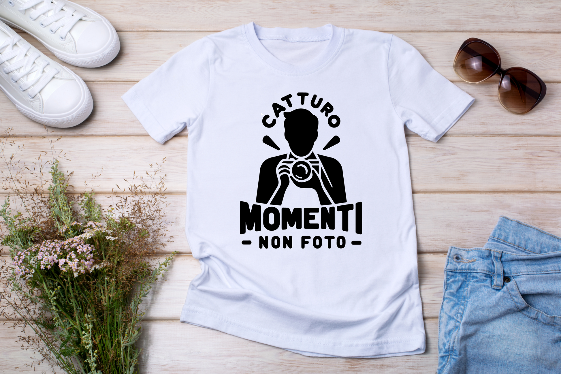 T-shirt bianca stesa con accessori
"T-shirt bianca con stampa nera 'Catturo Momenti, Non Foto', design con silhouette di un fotografo, posizionata su fondo in legno con occhiali da sole, jeans e fiori secchi."