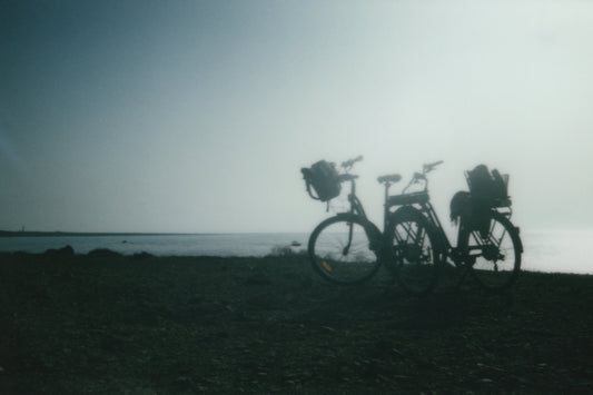 "Stampa Polaroid – Due biciclette appoggiate l'una all'altra in controluce, con il mare sullo sfondo, simbolo di viaggio e connessione."