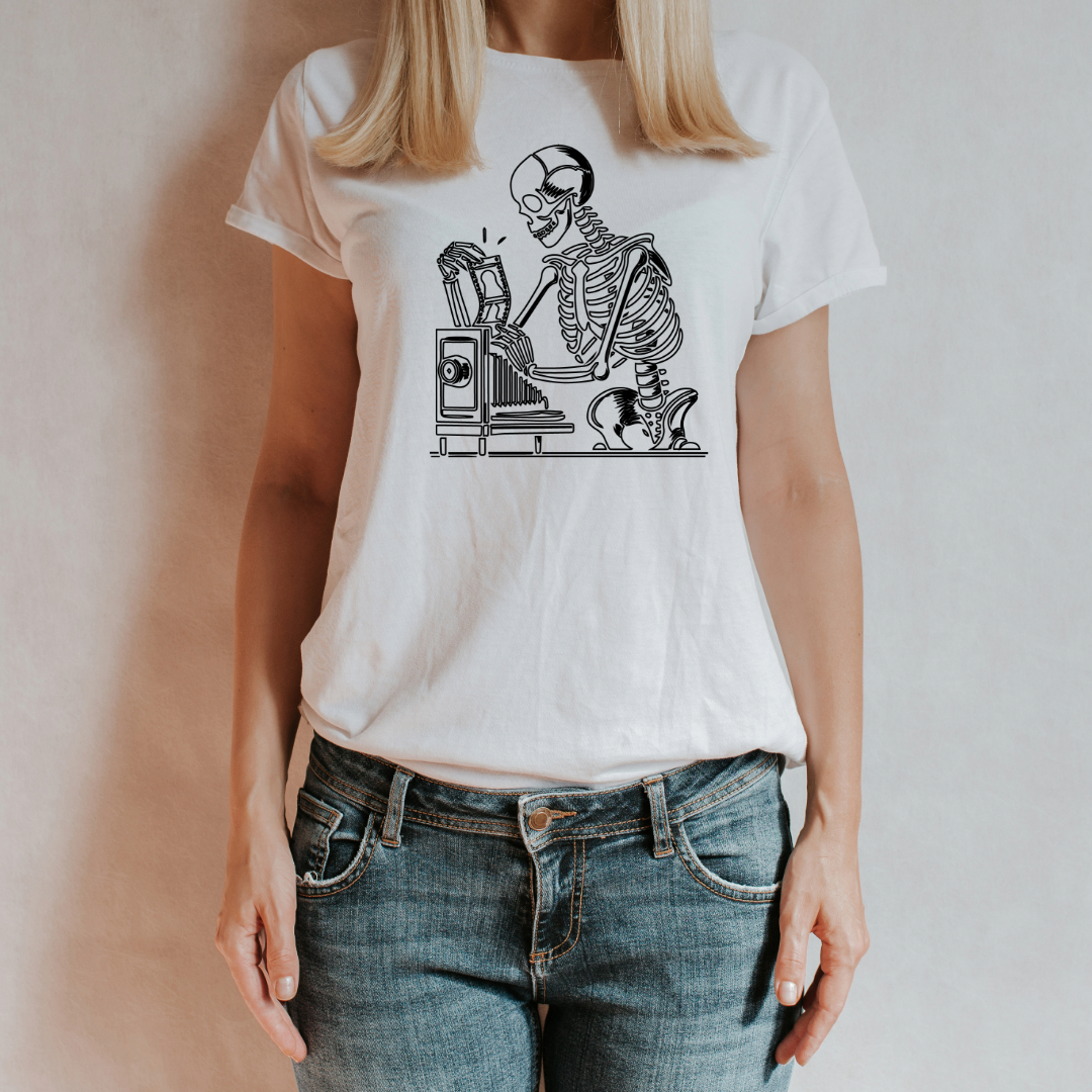 "T-shirt bianca 'Darkroom Bones' con stampa di uno scheletro che sviluppa foto in camera oscura, indossata da una donna con jeans, su sfondo neutro."