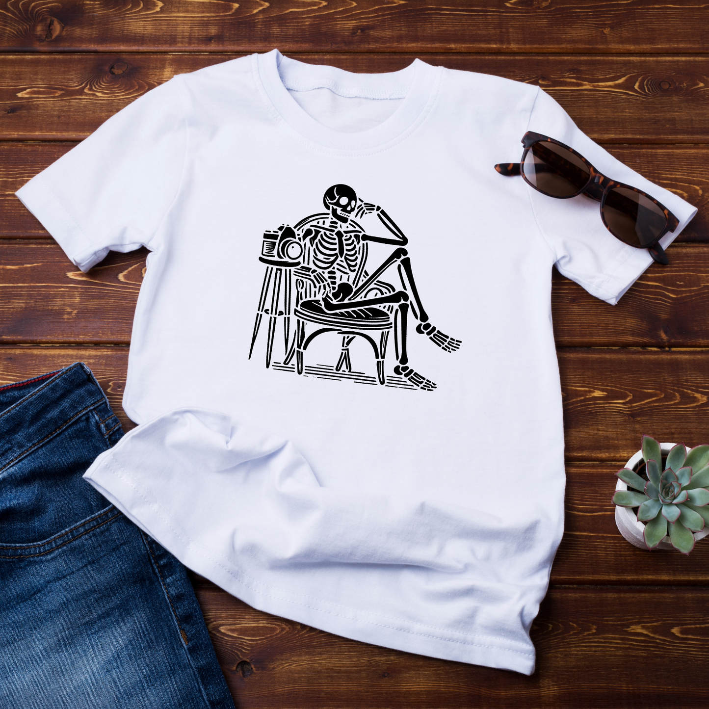 "T-shirt bianca con stampa artistica di uno scheletro seduto su una sedia con una macchina fotografica, in stile vintage minimalista. Disposta su fondo in legno con jeans e occhiali da sole come accessori."