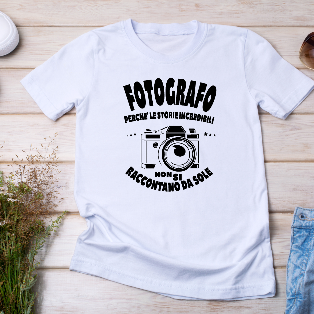 T-shirt bianca con design per fotografi, raffigurante una fotocamera vintage e una frase motivazionale. Disposta su una superficie in legno con occhiali da sole e decorazioni naturali.