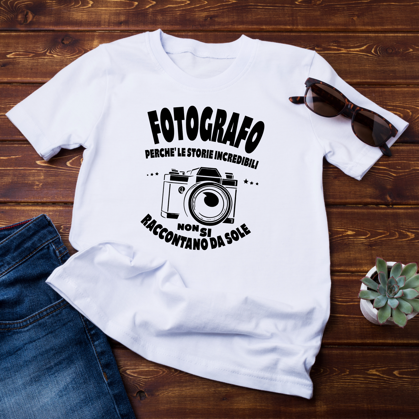 (maglietta flatlay su fondo in legno):
T-shirt bianca con stampa nera raffigurante una fotocamera vintage e la scritta "Fotografo – Perché le storie incredibili non si raccontano da sole", posata su una superficie in legno con accessori come occhiali da sole e jeans.