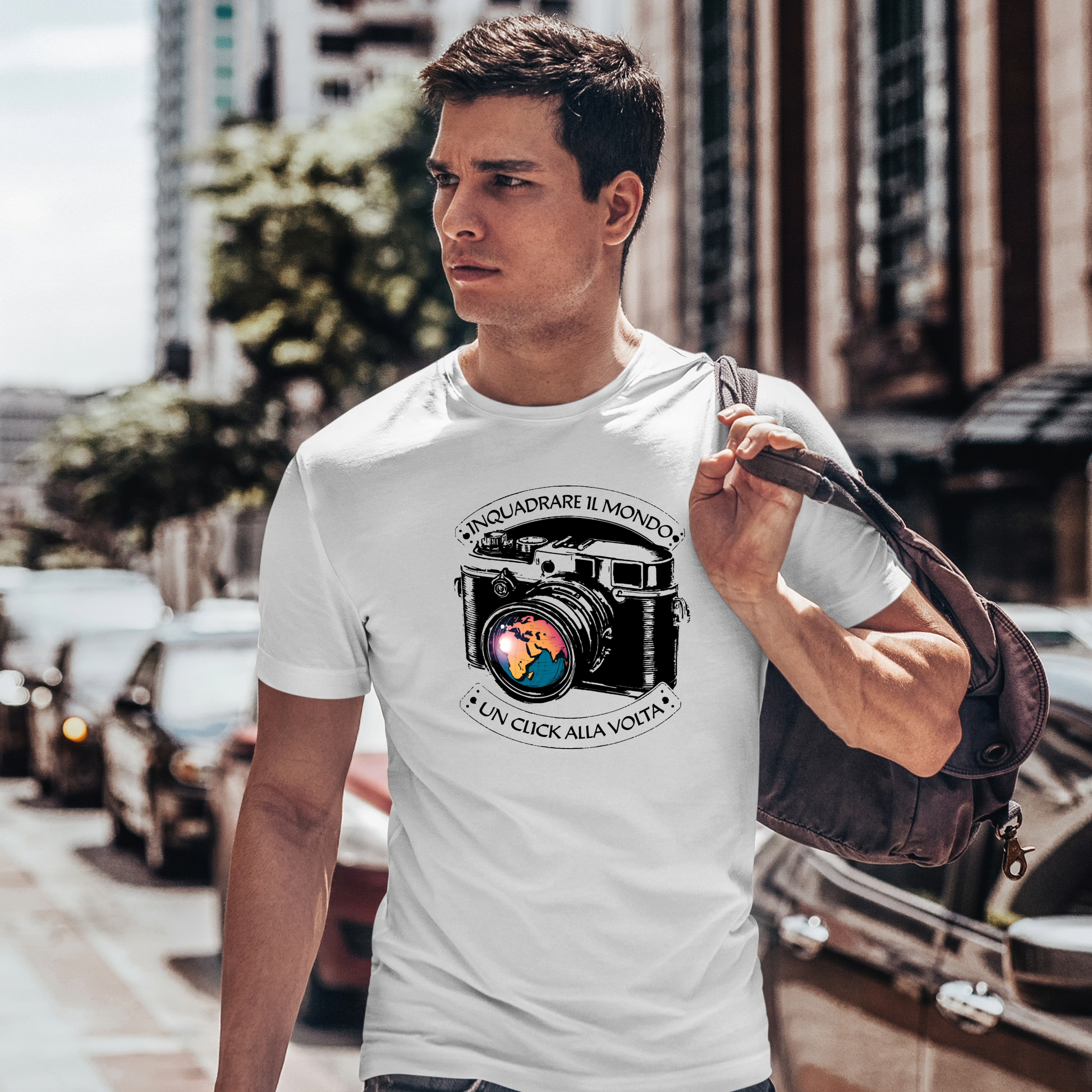 "Uomo che cammina in una strada di città indossando una t-shirt bianca con una grafica di una macchina fotografica vintage e la scritta 'Inquadrare il mondo, un click alla volta'. Porta una borsa a tracolla."