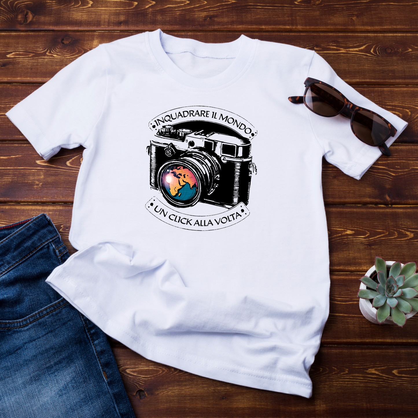 "T-shirt bianca con grafica di una fotocamera vintage e la Terra riflessa nell'obiettivo. Lo slogan 'Inquadrare il mondo, un click alla volta' avvolge il design. La maglietta è posata su una superficie in legno, accompagnata da occhiali da sole e jeans."