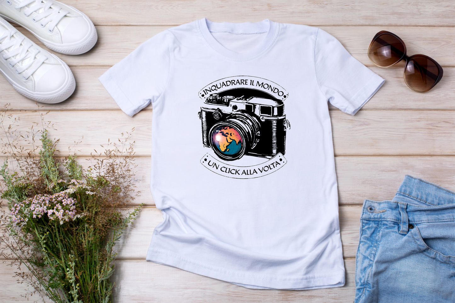 T-shirt bianca stesa con accessori
"T-shirt bianca 'Inquadrare il Mondo' con stampa di una fotocamera vintage e il mondo riflesso nell’obiettivo, posizionata su fondo in legno con occhiali da sole, jeans e fiori secchi