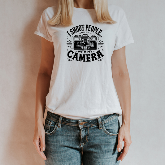 (Indossata Donna, posa su sfondo neutro): Donna con t-shirt bianca "I Shoot People with My Camera", look casual con jeans. Un capo ironico e di tendenza per fotografi e creativi.