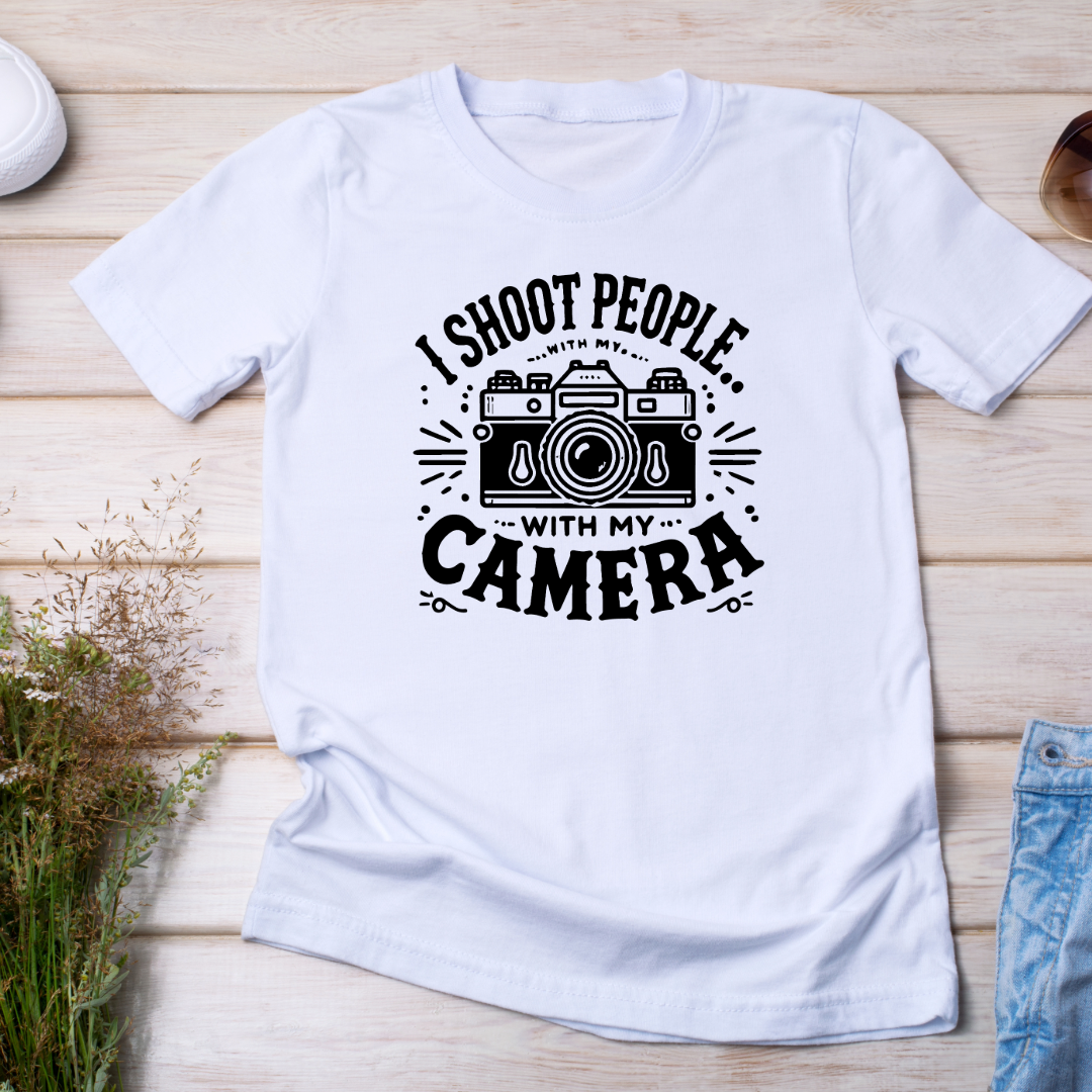 (T-shirt distesa su sfondo chiaro con jeans e occhiali): Maglietta bianca con stampa "I Shoot People with My Camera" affiancata da jeans e occhiali da sole. Ideale per outfit casual e amanti della fotografia