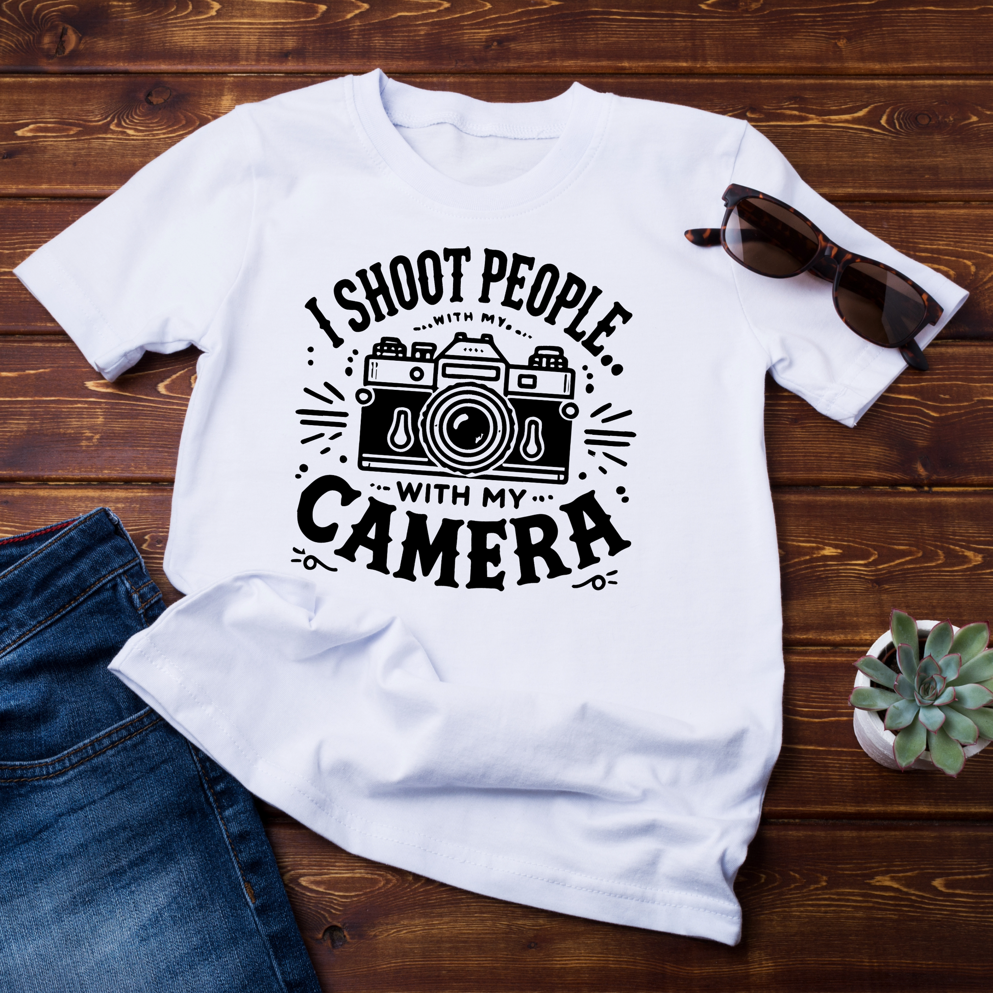 (Maglietta piegata su sfondo legno con occhiali da sole): T-shirt bianca "I Shoot People with My Camera" posata su superficie in legno, con occhiali da sole e pianta. Perfetta per fotografi che vogliono esprimere la propria passione con stile.