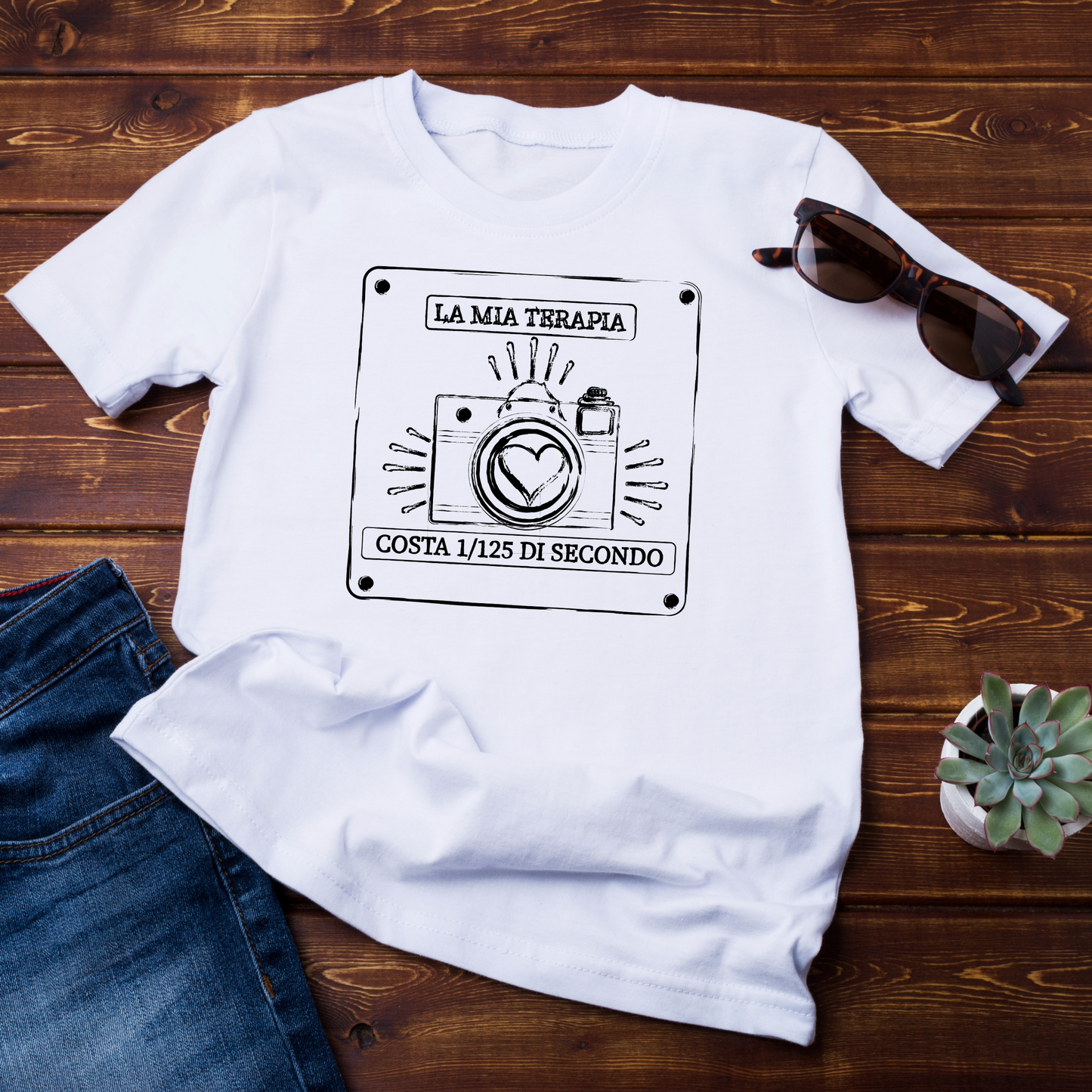 "T-shirt bianca con grafica fotografica e scritta motivazionale, posata su un fondo in legno con occhiali da sole e accessori."