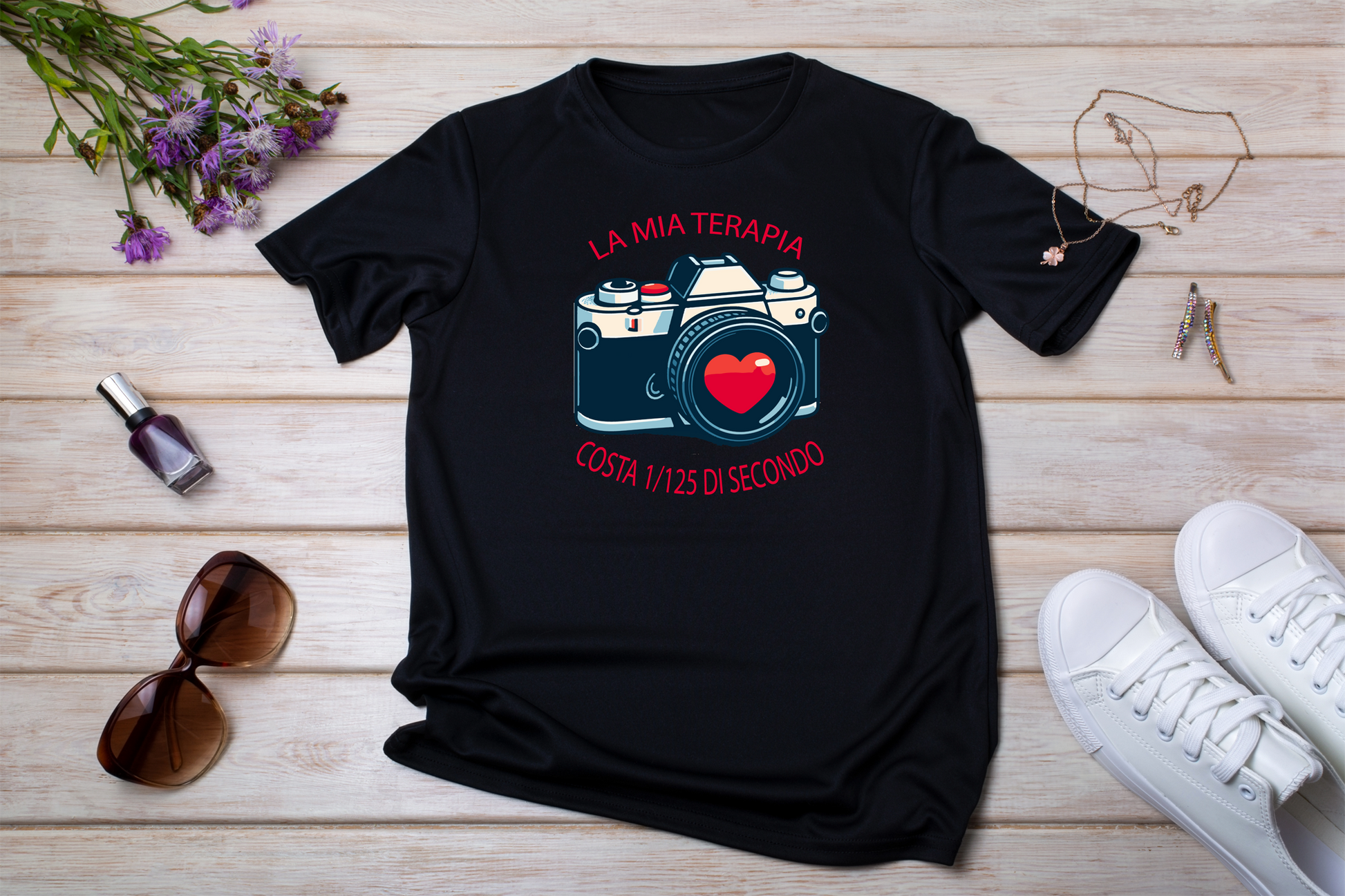 T-shirt nera stesa con accessori
"T-shirt nera 'La Mia Terapia' con stampa di una fotocamera vintage e cuore rosso nell'obiettivo, disposta su fondo in legno con occhiali da sole, fiori e gioielli