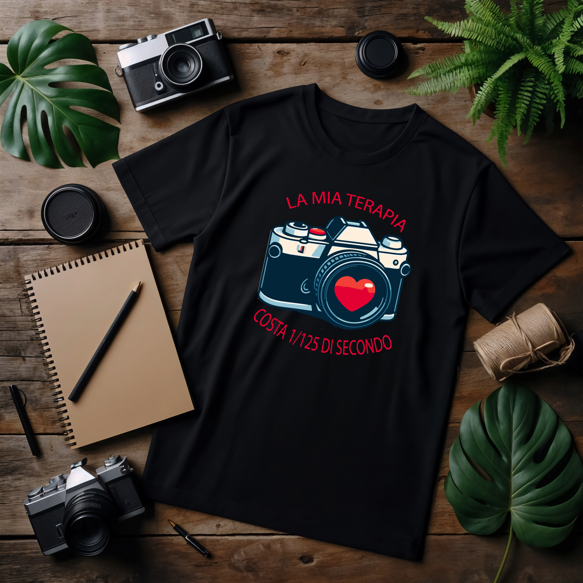 t-shirt in piano):
T-shirt nera con il design "Ama, Sorridi, Scatta", posizionata su un tavolo di legno con oggetti fotografici intorno. Il design è composto da un cuore verde, un volto sorridente e una macchina fotografica rossa, richiamando i colori della bandiera italiana.