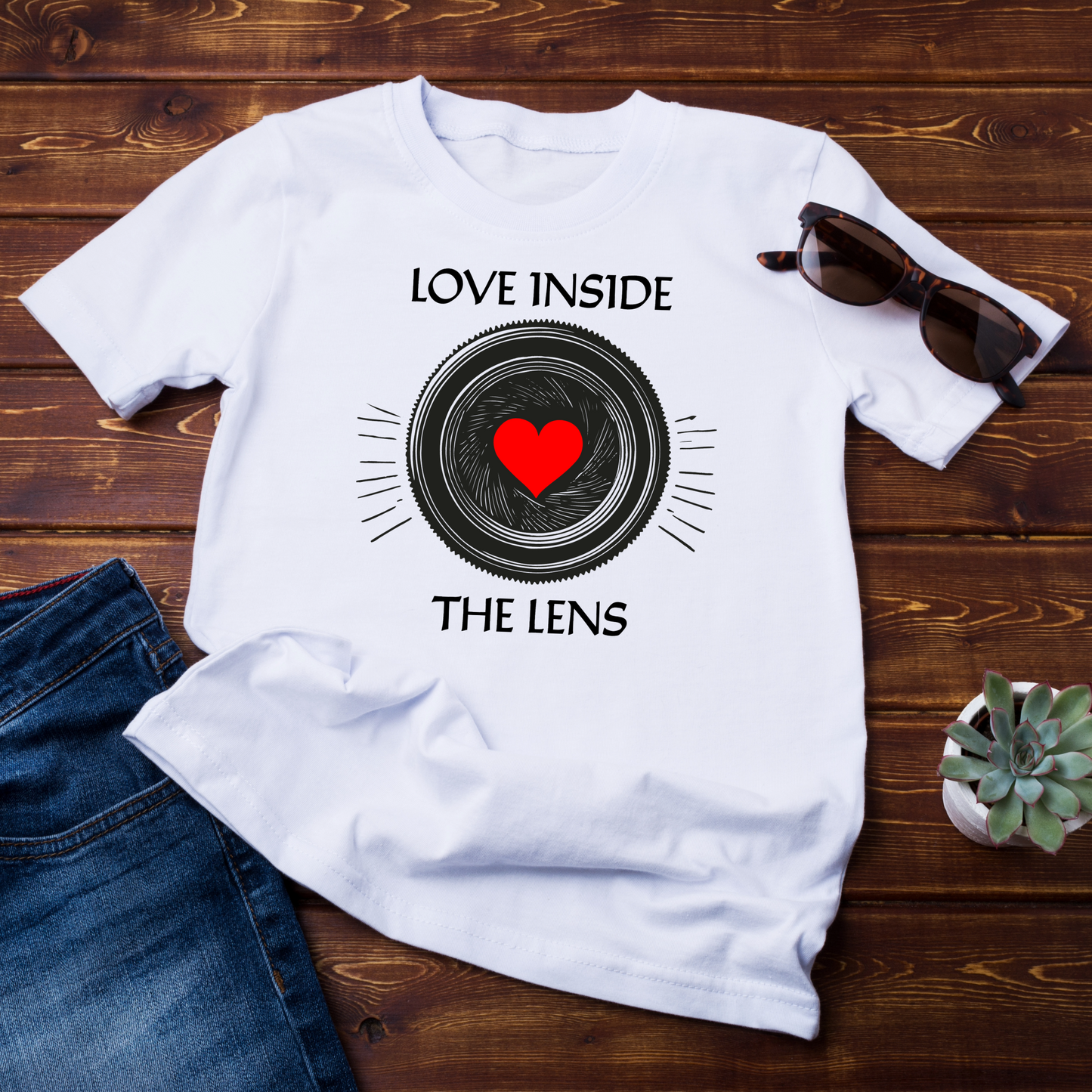 maglietta piegata su fondo in legno: T-shirt bianca "Love Inside The Lens" con design di un obiettivo fotografico e cuore rosso al centro. Perfetta per fotografi e appassionati di fotografia