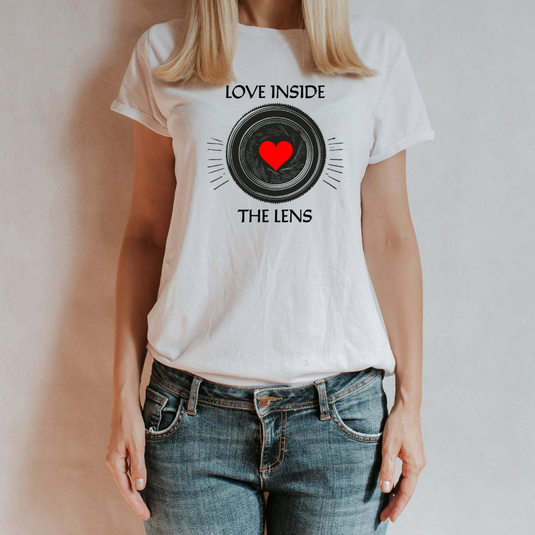 T-shirt bianca con stampa 'Love Inside the Lens', raffigurante un obiettivo fotografico con un cuore rosso al centro. Indossata da una donna con jeans, su sfondo neutro