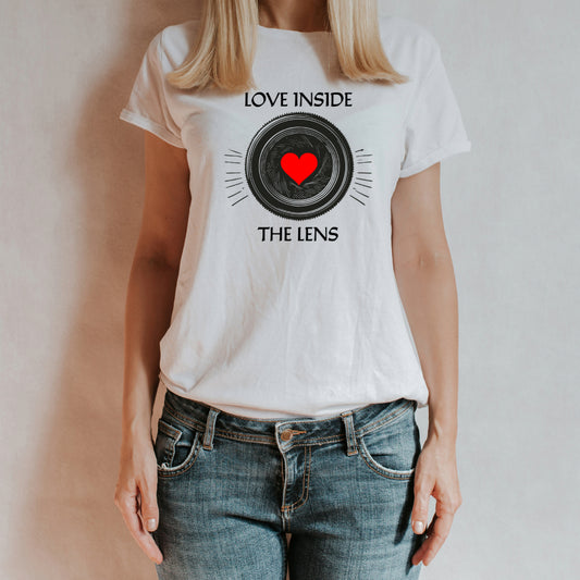 T-shirt bianca con stampa 'Love Inside the Lens', raffigurante un obiettivo fotografico con un cuore rosso al centro. Indossata da una donna con jeans, su sfondo neutro