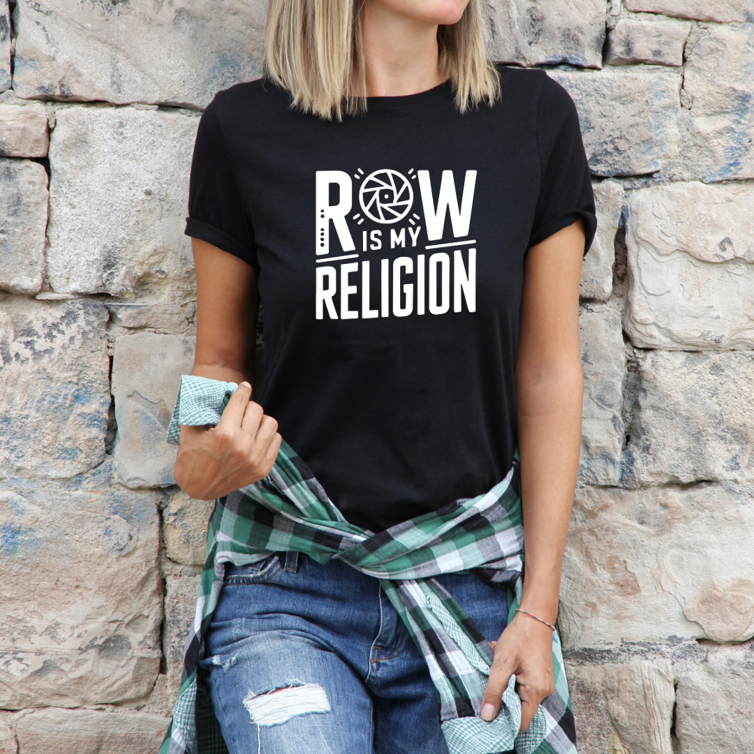 Maglietta "RAW is My Religion" in versione donna, indossata da una modella contro un muro di pietra, abbinata a jeans e camicia a quadri.