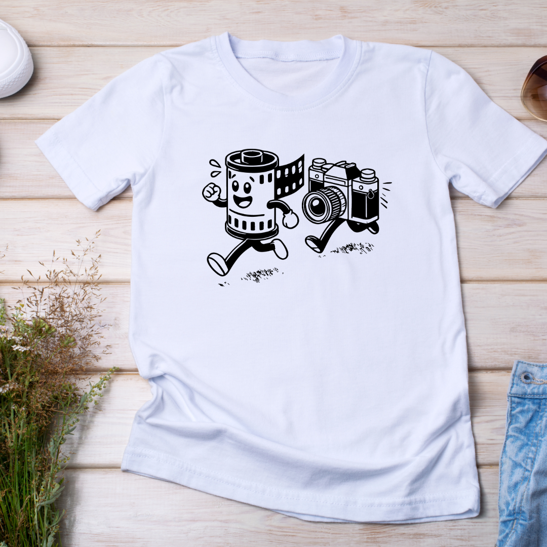 T-shirt bianca distesa su una superficie di legno chiaro, con il design "Run & Shoot" che raffigura un rullino e una fotocamera vintage in corsa.