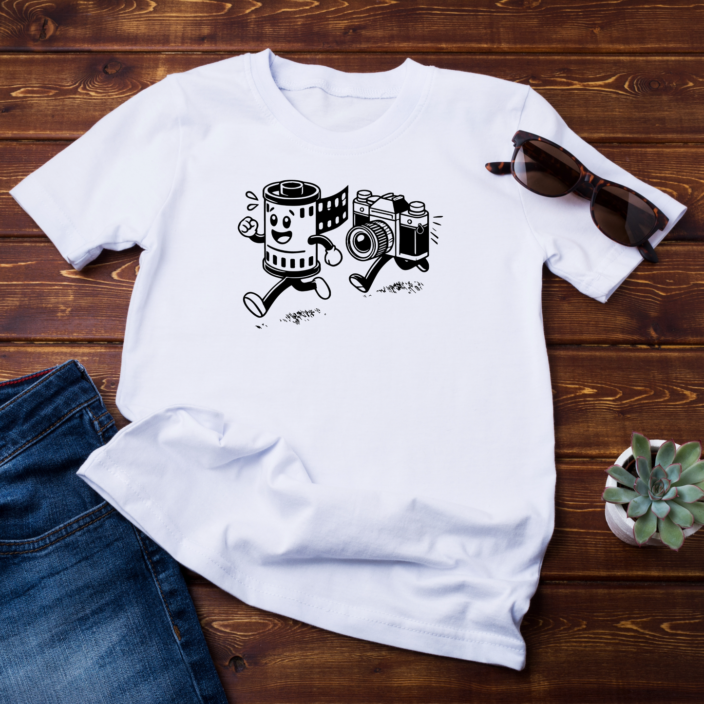 T-shirt bianca appoggiata su un piano di legno scuro, accompagnata da accessori casual come occhiali da sole e una piantina. Il design in evidenza raffigura un rullino e una fotocamera vintage in corsa.