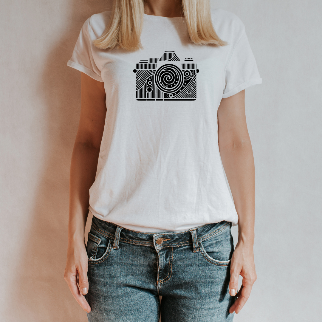 T-shirt bianca 'Spiral Lens' con grafica astratta di una fotocamera e dettagli geometrici, indossata da una donna con jeans, su sfondo neutro