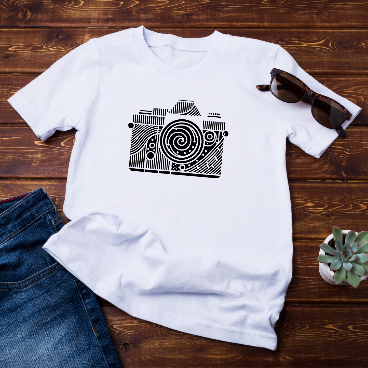 "T-shirt bianca con stampa artistica di una fotocamera decorata con motivi geometrici e una spirale centrale. La maglietta è posata su una superficie in legno con occhiali da sole e jeans come accessori."