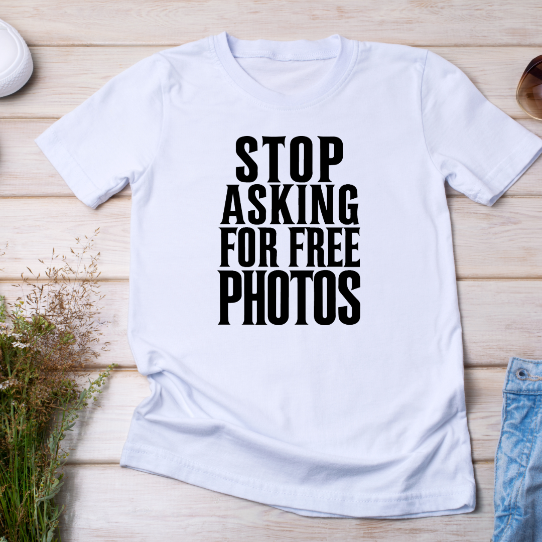 (T-shirt bianca piegata su sfondo in legno con accessori fotografici):
"Maglietta bianca 'Stop Asking for Free Photos' su fondo in legno, ideale per fotografi che vogliono esprimere con stile il valore della loro arte."