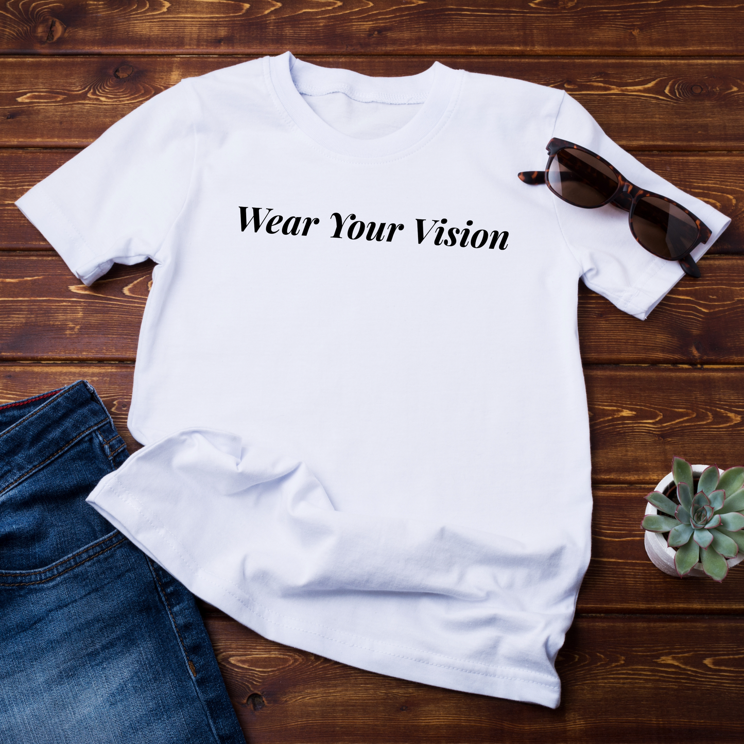 (flat lay su sfondo scuro)
"T-shirt bianca con la scritta 'Wear Your Vision' in nero, disposta su un fondo di legno scuro con occhiali da sole, jeans e una pianta decorativa."