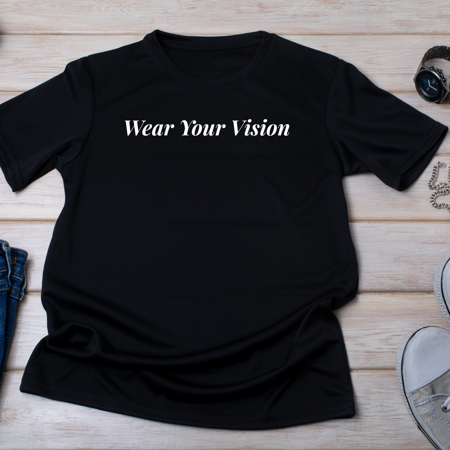 (flat lay su sfondo chiaro)
"T-shirt nera con la scritta 'Wear Your Vision' in carattere elegante, posizionata su un fondo di legno chiaro con dettagli di moda come orologio, gioielli e scarpe bianche.