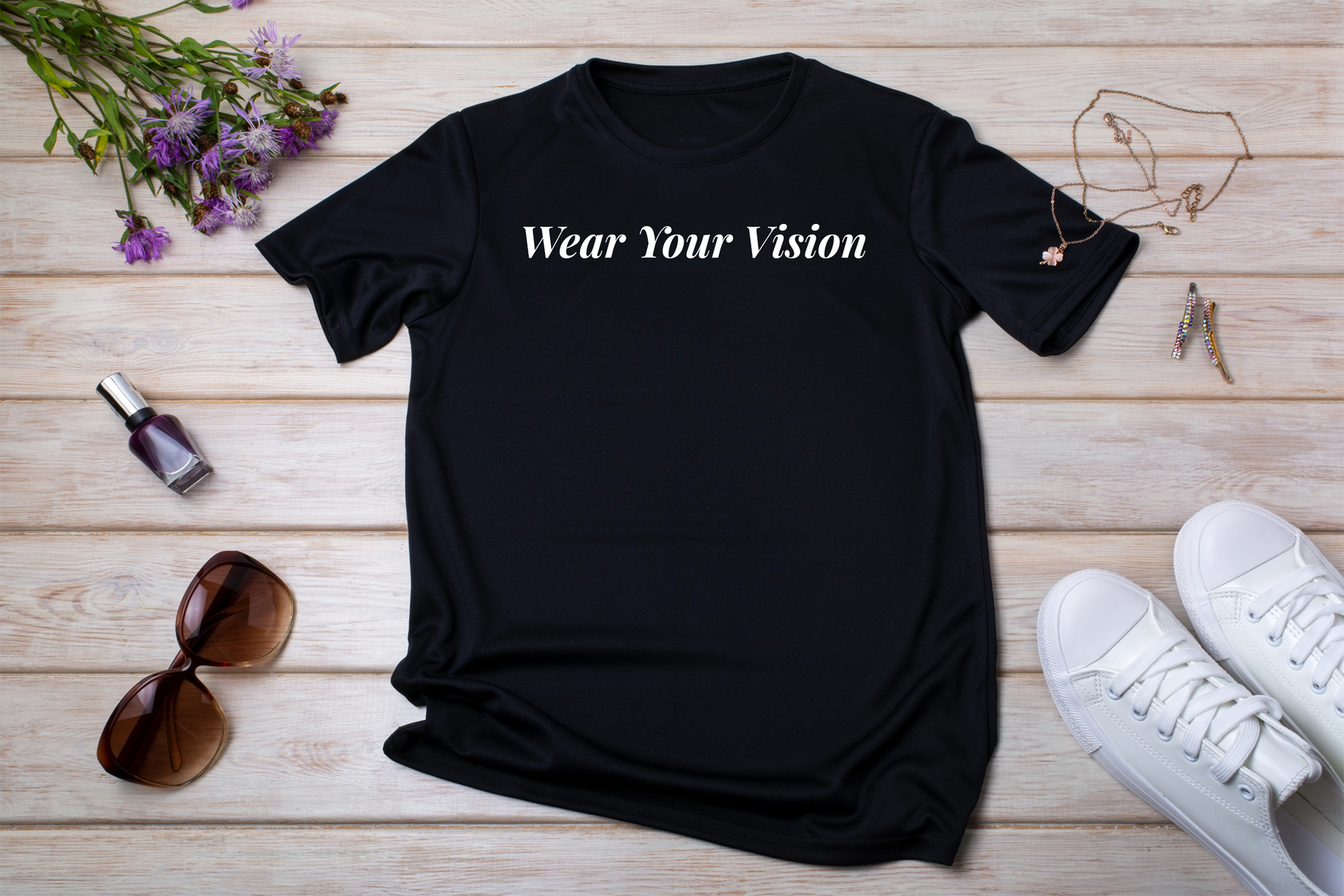 T-shirt nera stesa con accessori
"T-shirt nera con la scritta 'Wear Your Vision' in bianco, disposta su fondo in legno accanto a occhiali da sole, gioielli e sneakers bianche."