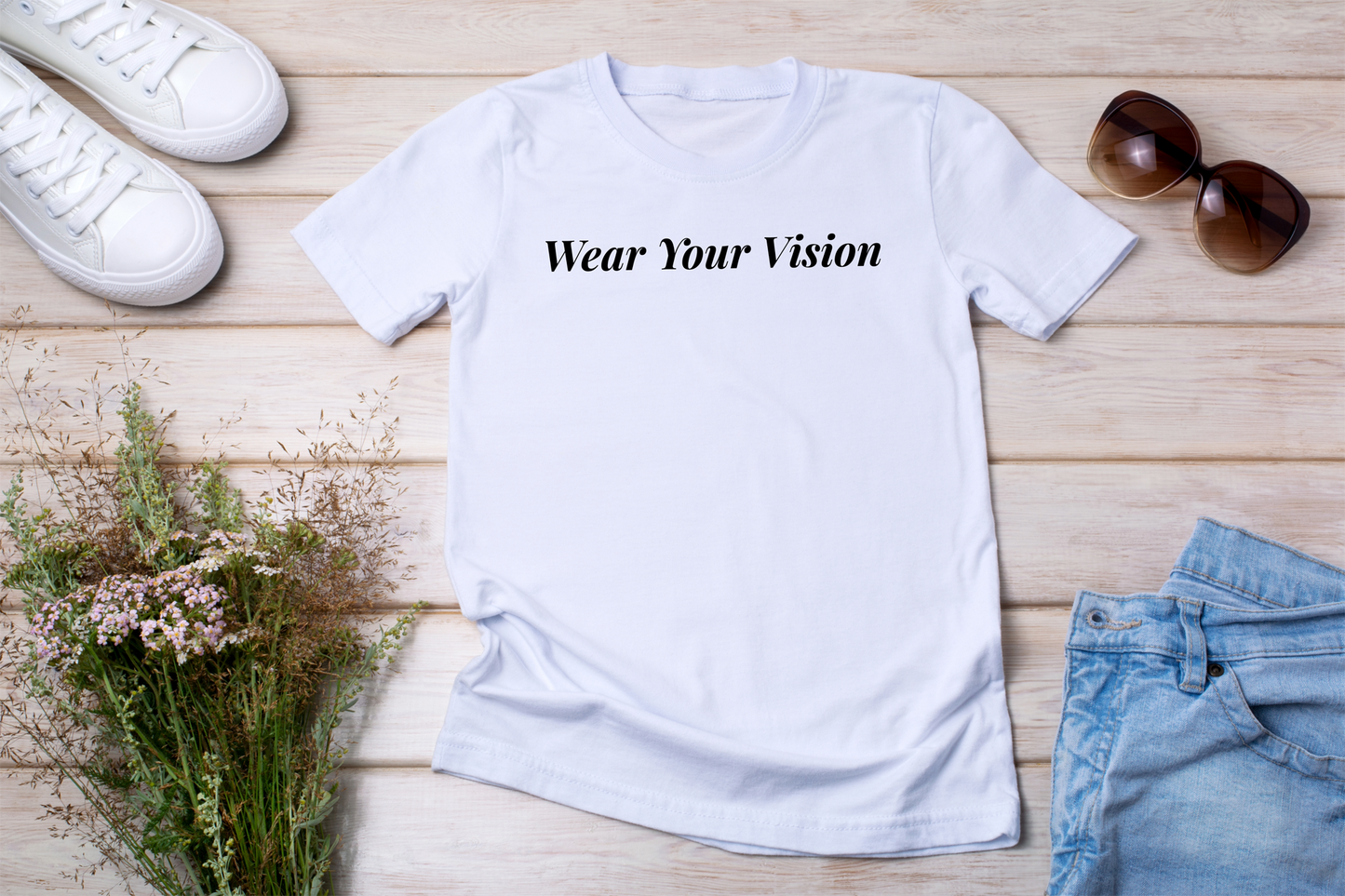 1️⃣ T-shirt bianca stesa con accessori
"T-shirt bianca con la scritta 'Wear Your Vision' in nero, posizionata su fondo in legno con occhiali da sole, jeans e fiori secchi."