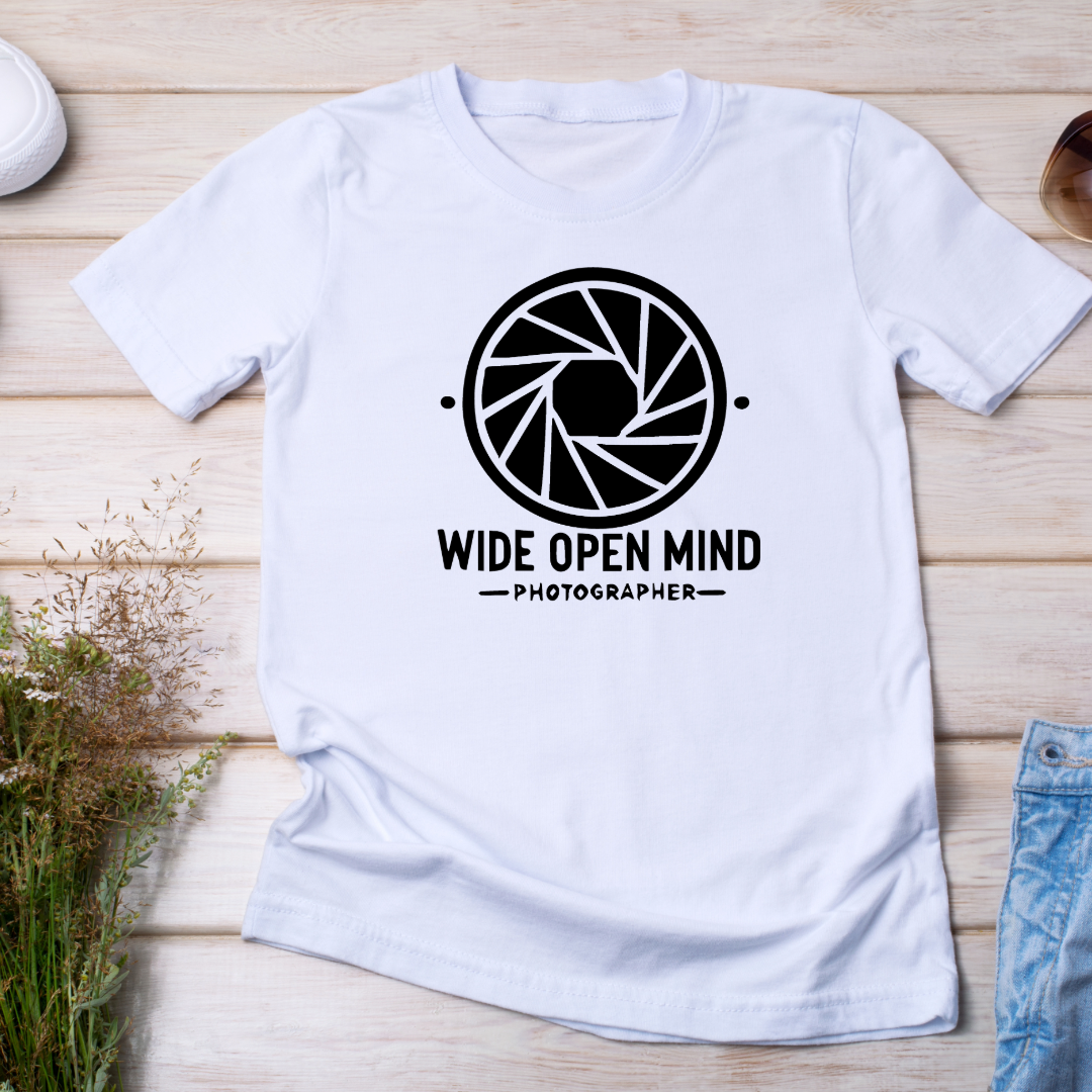 T-shirt bianca "Wide Open Mind" posata su una superficie in legno con occhiali da sole e piantina, evidenziando la qualità del tessuto.